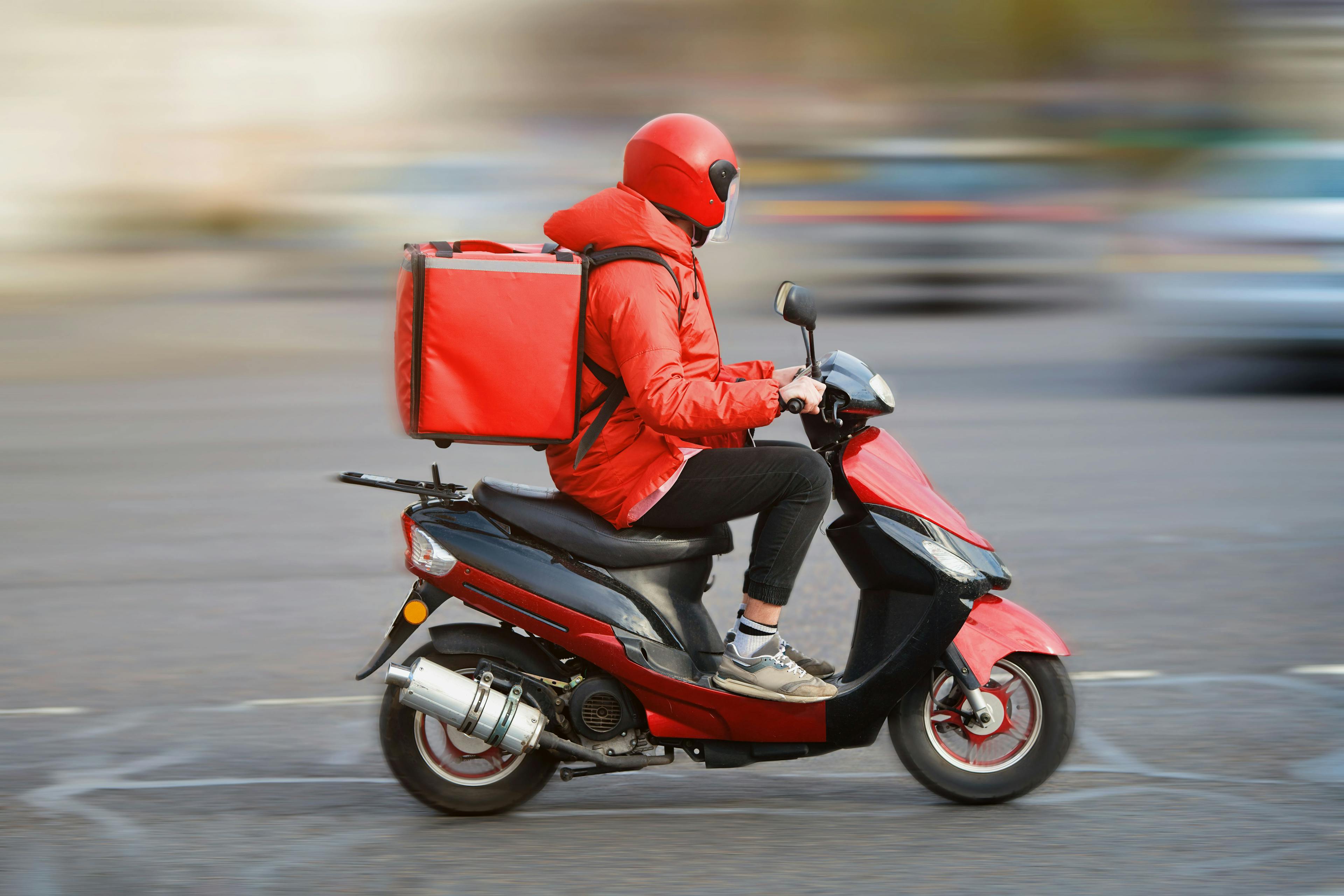 A man wearing a red jacket rides a scooter, transporting a box for delivery.