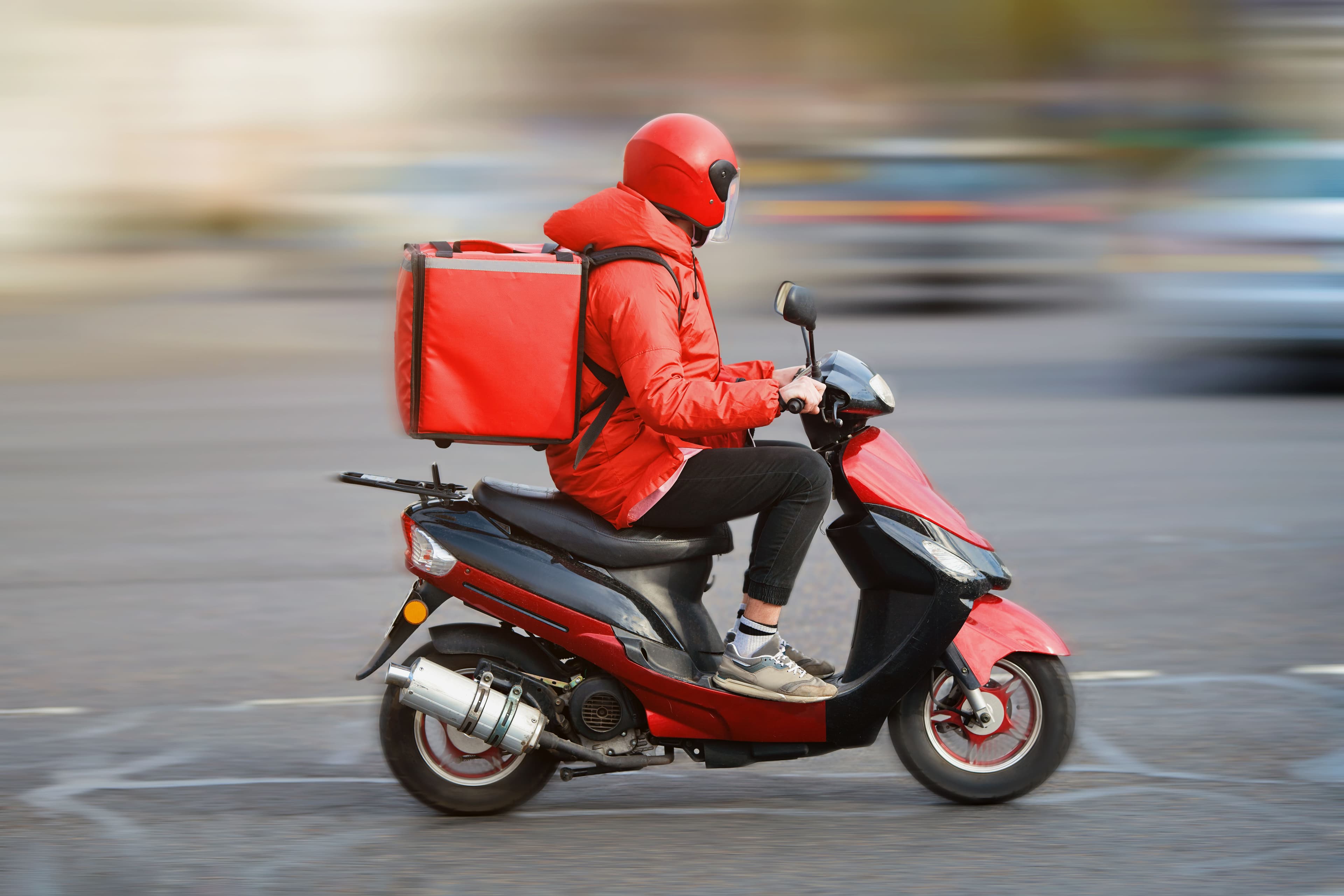 A man wearing a red jacket rides a scooter, transporting a box for delivery.