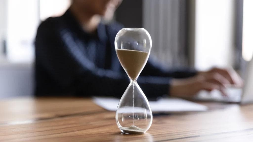 An individual engrossed in work at a desk, accompanied by a laptop and an hourglass as a reminder of time management.