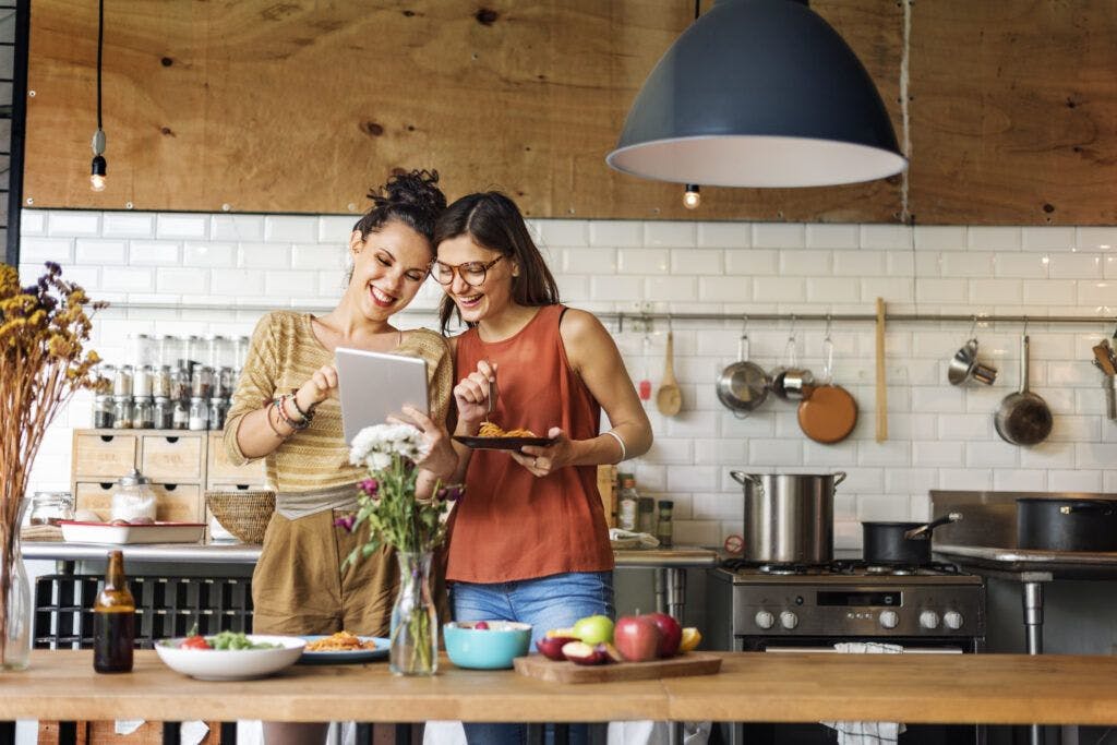 Two women in a kitchen, focused on a tablet displaying a menu.