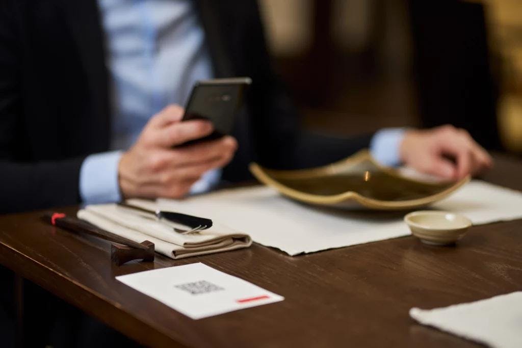 A man in formal attire sitting at a table, focused on his phone, with a visible menu on the table.
