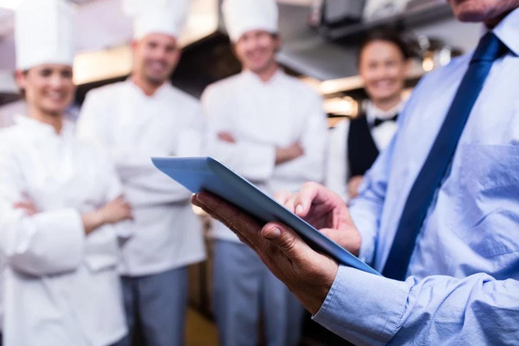 A chef wearing a hat holds a tablet, possibly using it for recipes or managing kitchen operations.